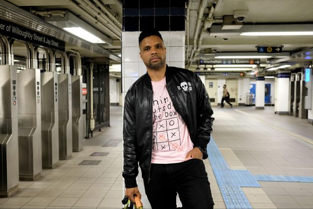 A man wearing a black jacket and pink shirt stands in a subway station. after the tuurnstiles